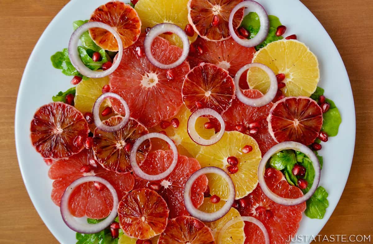 A salad of sliced oranges, blood oranges and grapefruits, red onion slices, arugula and pomegranate seeds on a white serving platter.