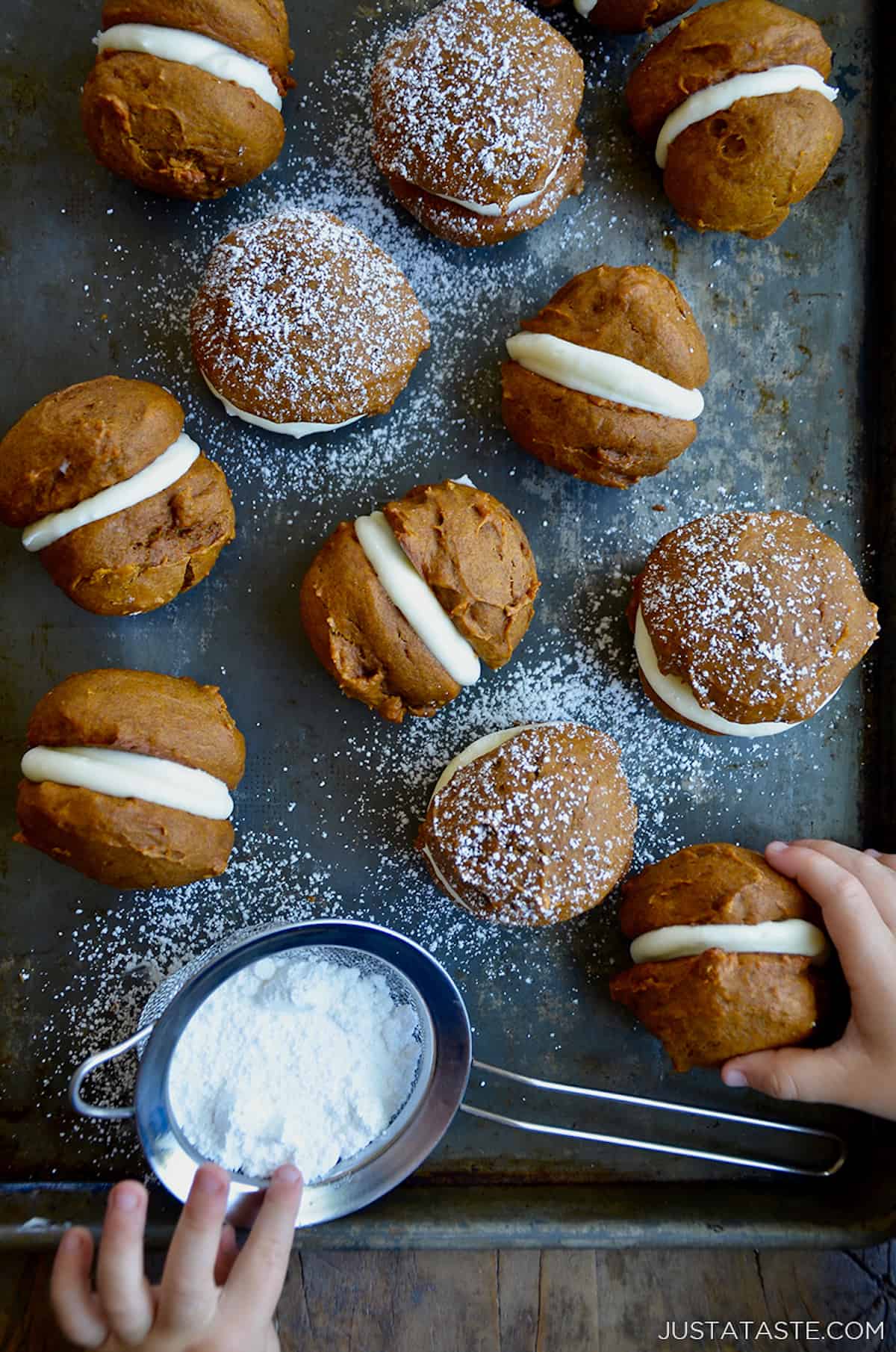 A sheet tray with pumpkin whoopie pies dusted with powdered sugar. A small fine mesh sieve with more powdered sugar sits off to the side and a small child's hands are grabbing a whoopie pie.
