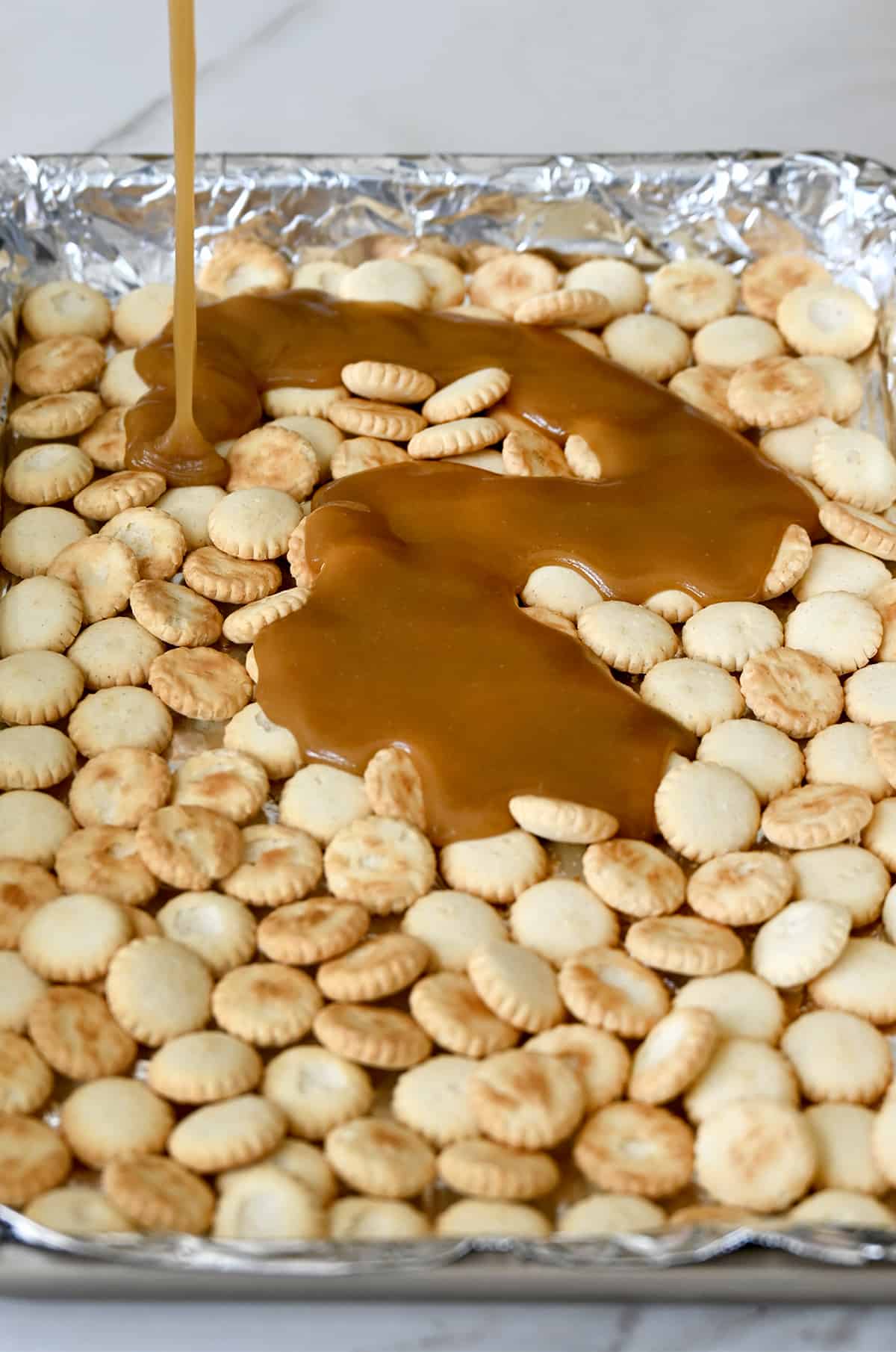 Toffee sauce being poured atop oyster crackers on a foil-lined baking sheet.