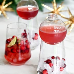 Three cocktail glasses containing ice, fresh cranberries and cranberry margarita mix.