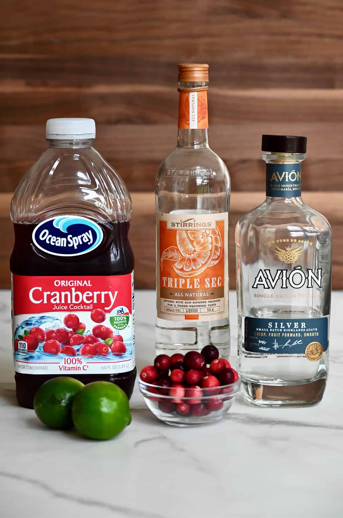 A jug of cranberry juice next to two limes, a bottle of Triple Sec, a bottle of silver tequila and a small bowl filled with fresh cranberries.