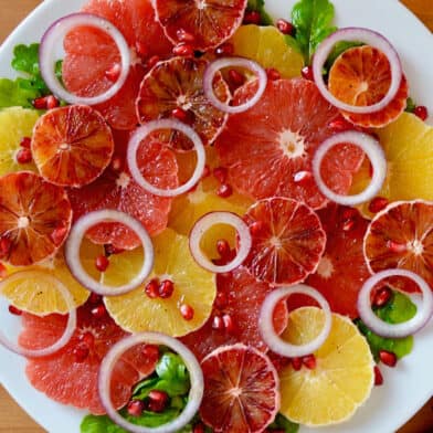 A salad comprised of sliced oranges, blood oranges and grapefruits, red onion slices, arugula and pomegranate seeds on a white serving platter. Salad tongs sit beside the platter.