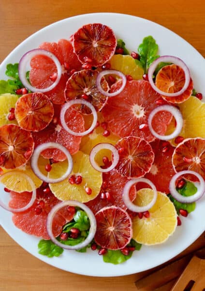 A salad comprised of sliced oranges, blood oranges and grapefruits, red onion slices, arugula and pomegranate seeds on a white serving platter. Salad tongs sit beside the platter.