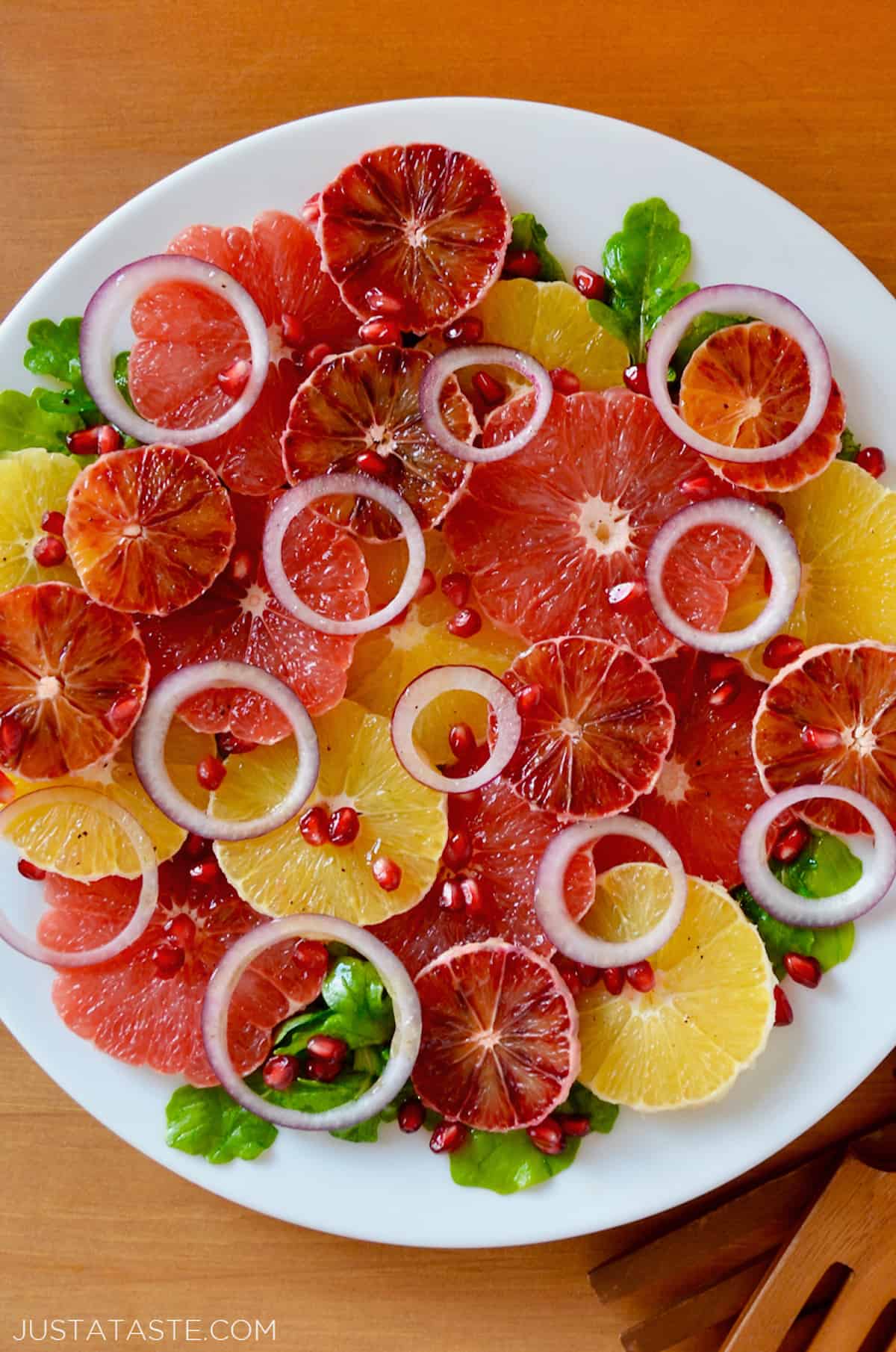 A salad comprised of sliced oranges, blood oranges and grapefruits, red onion slices, arugula and pomegranate seeds on a white serving platter. Salad tongs sit beside the platter.