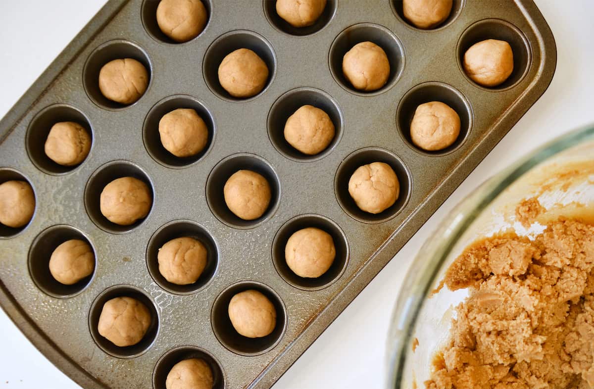 Balls of cookie dough in a mini muffin tin. A glass bowl containing more cooke dough is beside the tin.