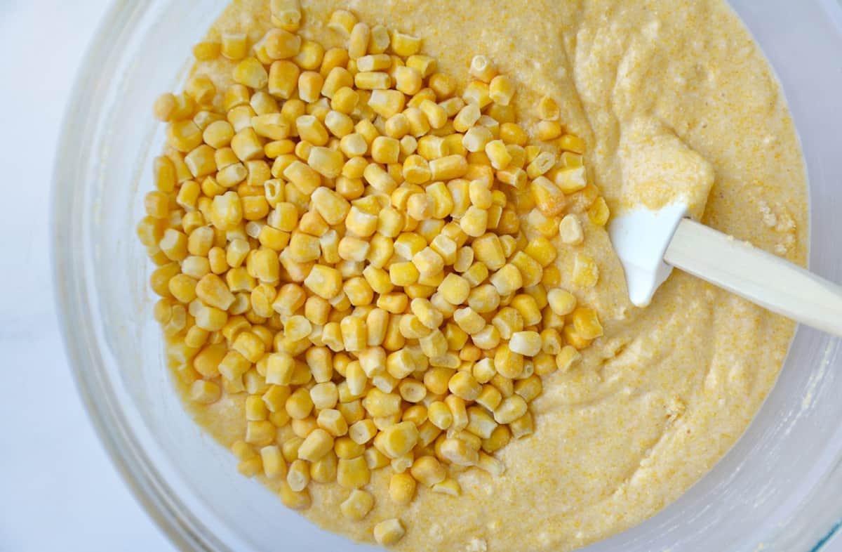 Cornbread batter in a glass mixing bowl with corn kernels on top of the batter and a rubber spatula propped against the side of of the bowl.