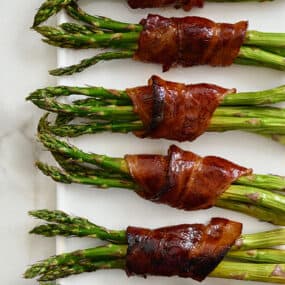 Six cooked bacon-wrapped asparagus bundles on a white platter.