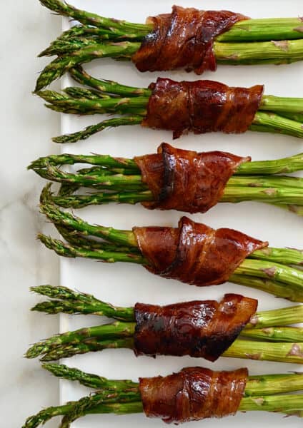 Six cooked bacon-wrapped asparagus bundles on a white platter.