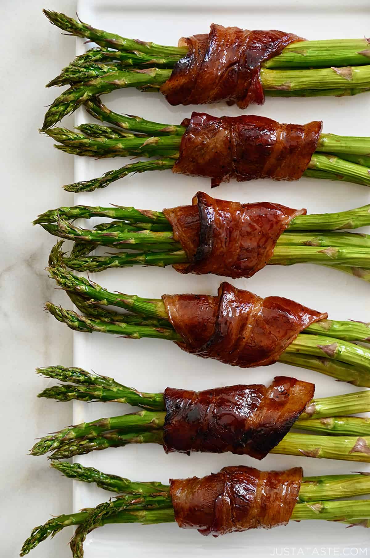 Six cooked bacon-wrapped asparagus bundles on a white platter.