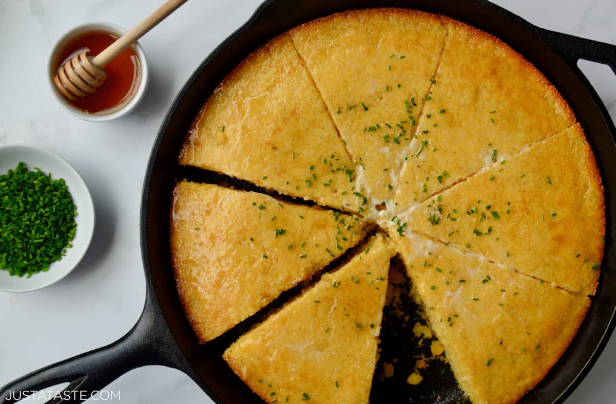 Baked, sliced cornbread in a cast iron skillet topped with butter, minced chives and honey. Small bowls of more minced chives and honey are beside the skillet.