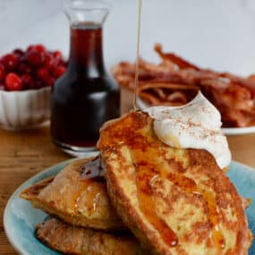 Maple syrup being poured onto a plate of three slices of French toast topped with whipped cream. A carafe of maple syrup, a plate of crispy bacon and a bowl of cranberries are behind the French toast.