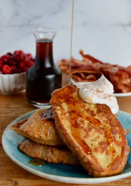 Maple syrup being poured onto a plate of three slices of French toast topped with whipped cream. A carafe of maple syrup, a plate of crispy bacon and a bowl of cranberries are behind the French toast.