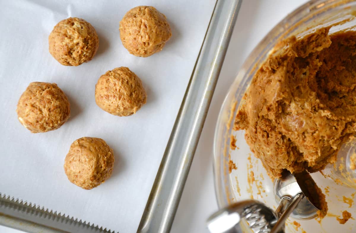 Five gingerbread cookie balls on a parchment-lined baking sheet. A glass bowl containing more dough and a portion scoop are beside the baking sheet.