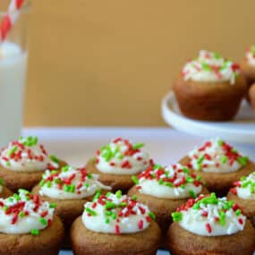 Gingerbread cookie cups topped with cream cheese frosting and sprinkles on a white platter. A glass of milk and a plate with more cookie cups sit behind the platter.