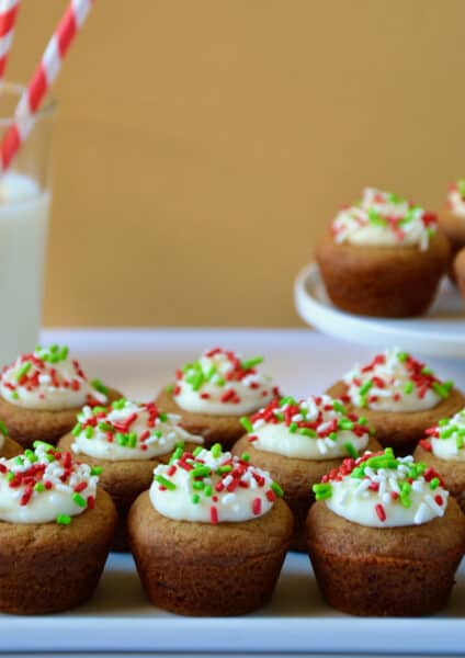 Gingerbread cookie cups topped with cream cheese frosting and sprinkles on a white platter. A glass of milk and a plate with more cookie cups sit behind the platter.