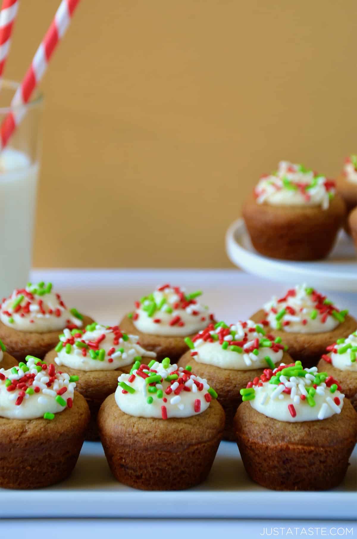 Gingerbread cookie cups topped with cream cheese frosting and sprinkles on a white platter. A glass of milk and a plate with more cookie cups sit behind the platter.
