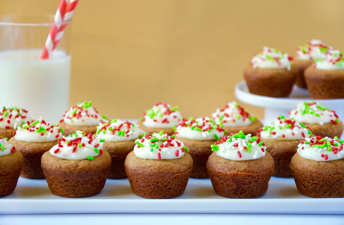 Gingerbread cookie cups topped with cream cheese frosting and green, white and red sprinkles on a white platter. A glass of milk and a plate with more cookie cups sit behind the platter.