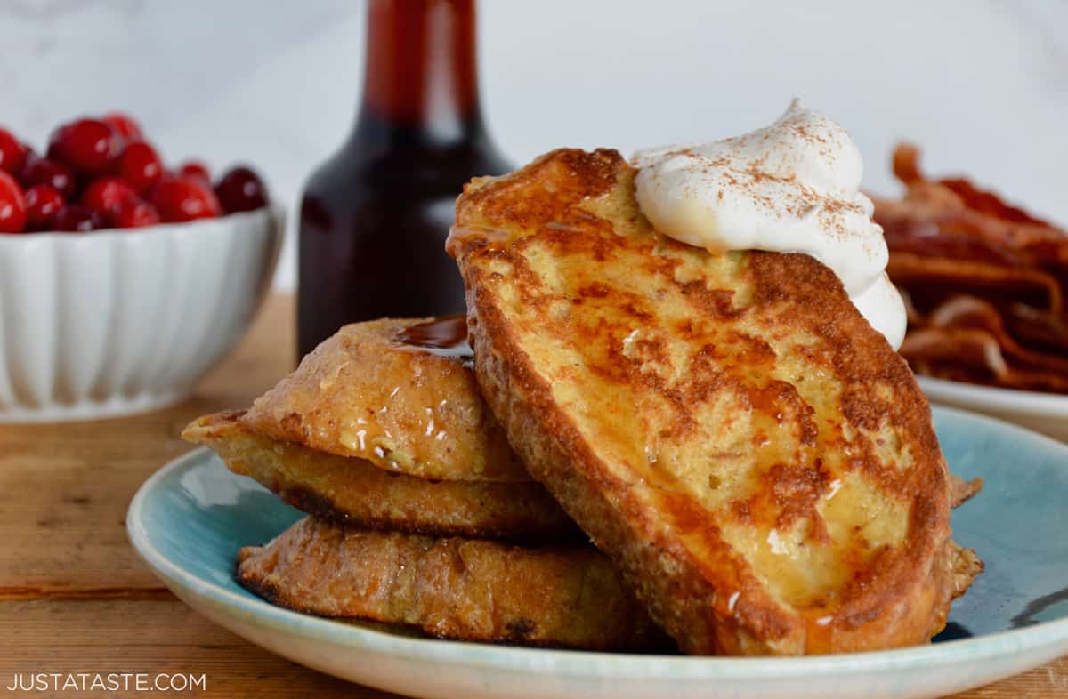 A plate of three slices of French toast topped with whipped cream. A carafe of maple syrup, a plate of crispy bacon and a bowl of cranberries are behind the French toast.