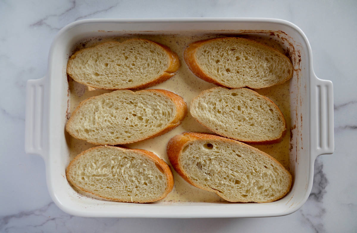 Six slices of bread soaking in custard in a rectangular baking dish.