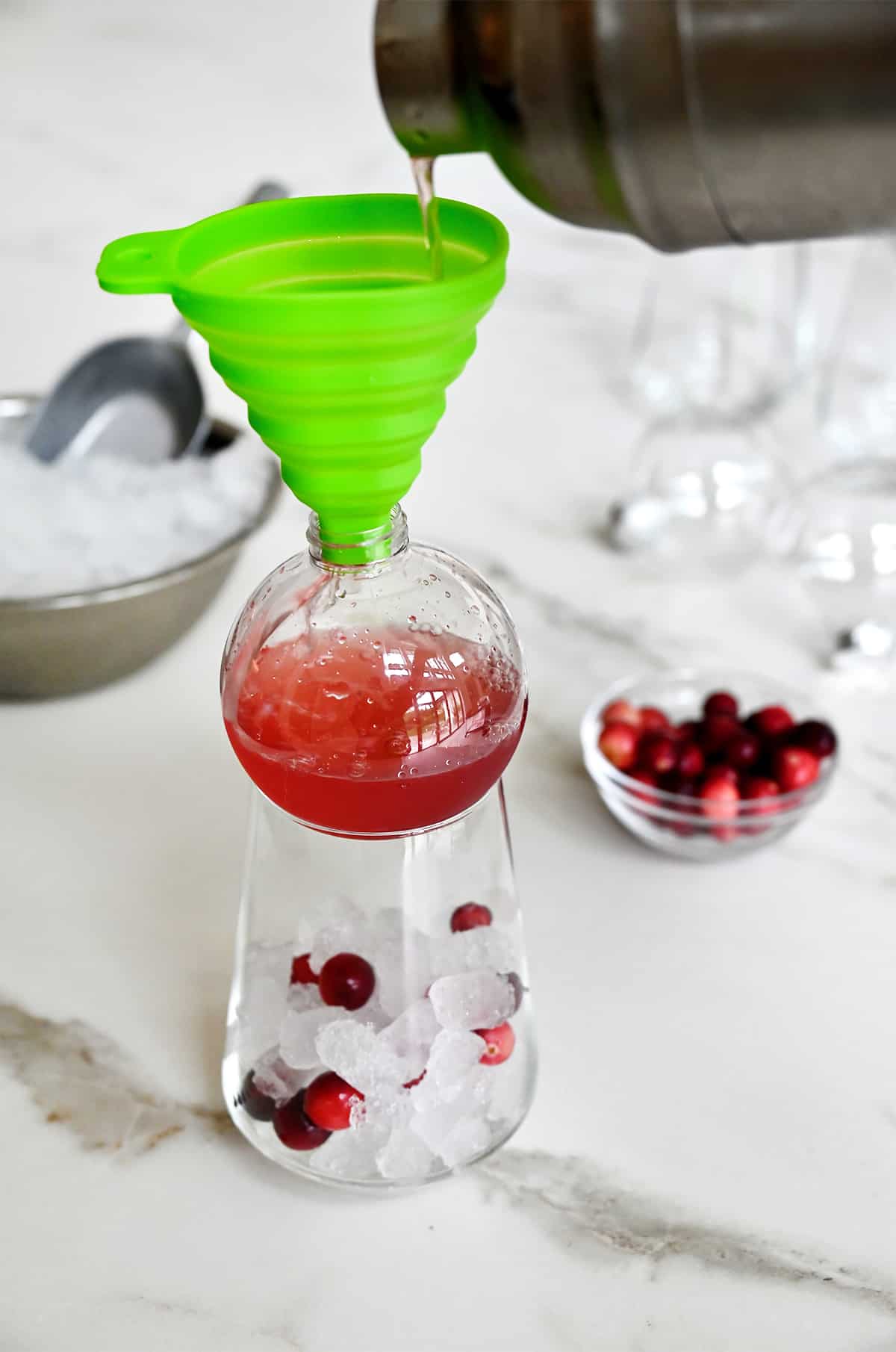 A cocktail shaker pours margarita mix into a funnel that's set in a clear ornament drinking ball atop a cocktail glass.