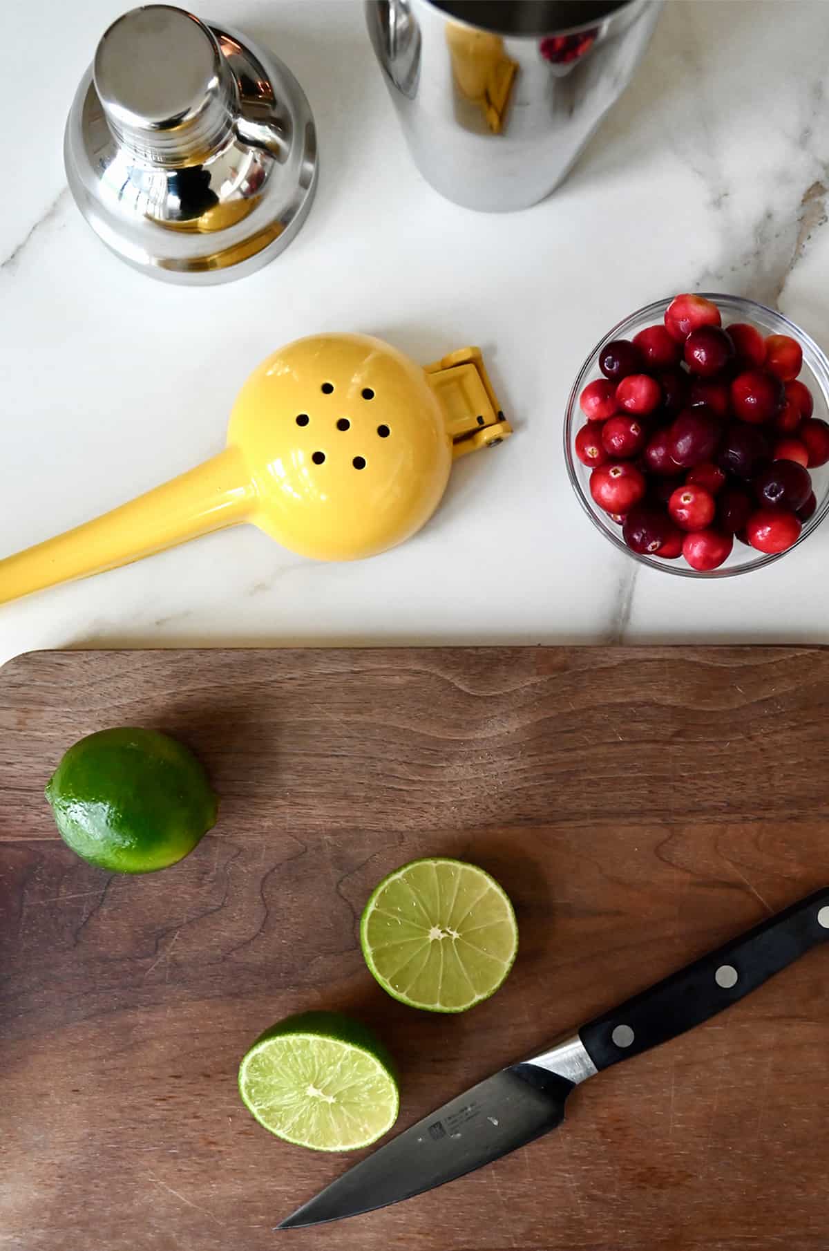 A halved lime on a cutting board with a sharp knife is next to a handheld juicer and a small bowl containing fresh cranberries.