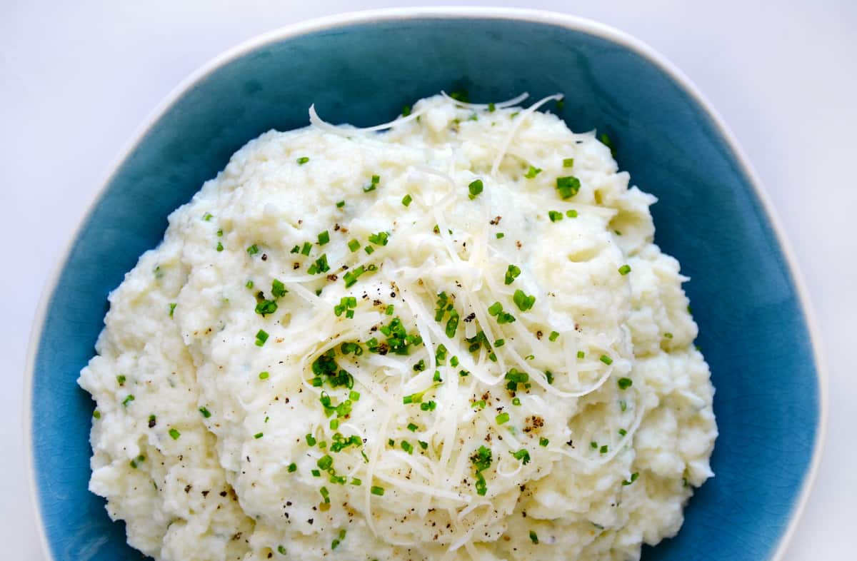 Mashed cauliflower topped with grated cheese, minced chives and black pepper in a blue bowl.