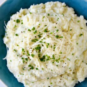 Mashed cauliflower topped with shredded cheese, finely minced chives and black pepper in a blue bowl.