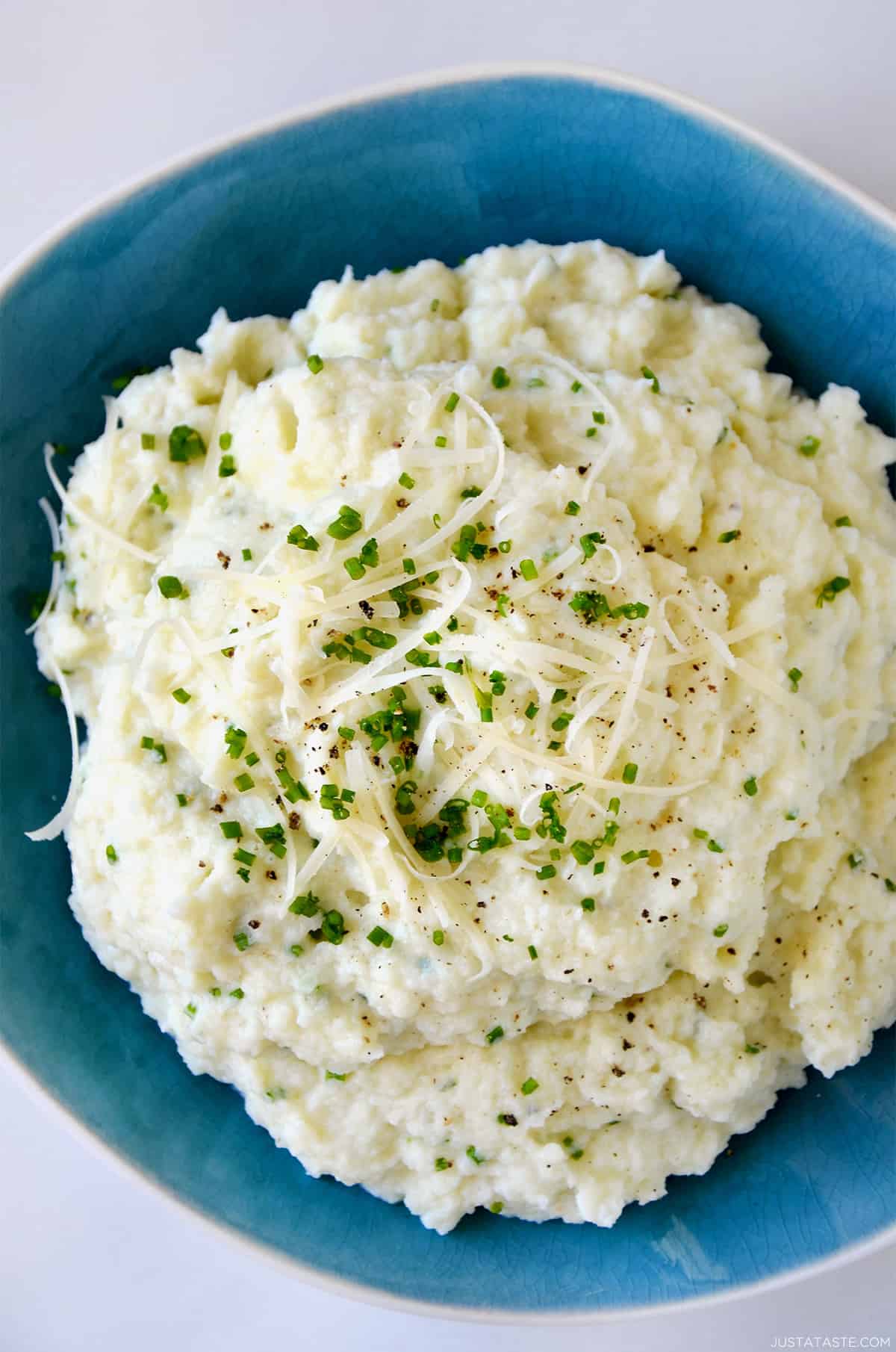 Mashed cauliflower topped with shredded cheese, finely minced chives and black pepper in a blue bowl.