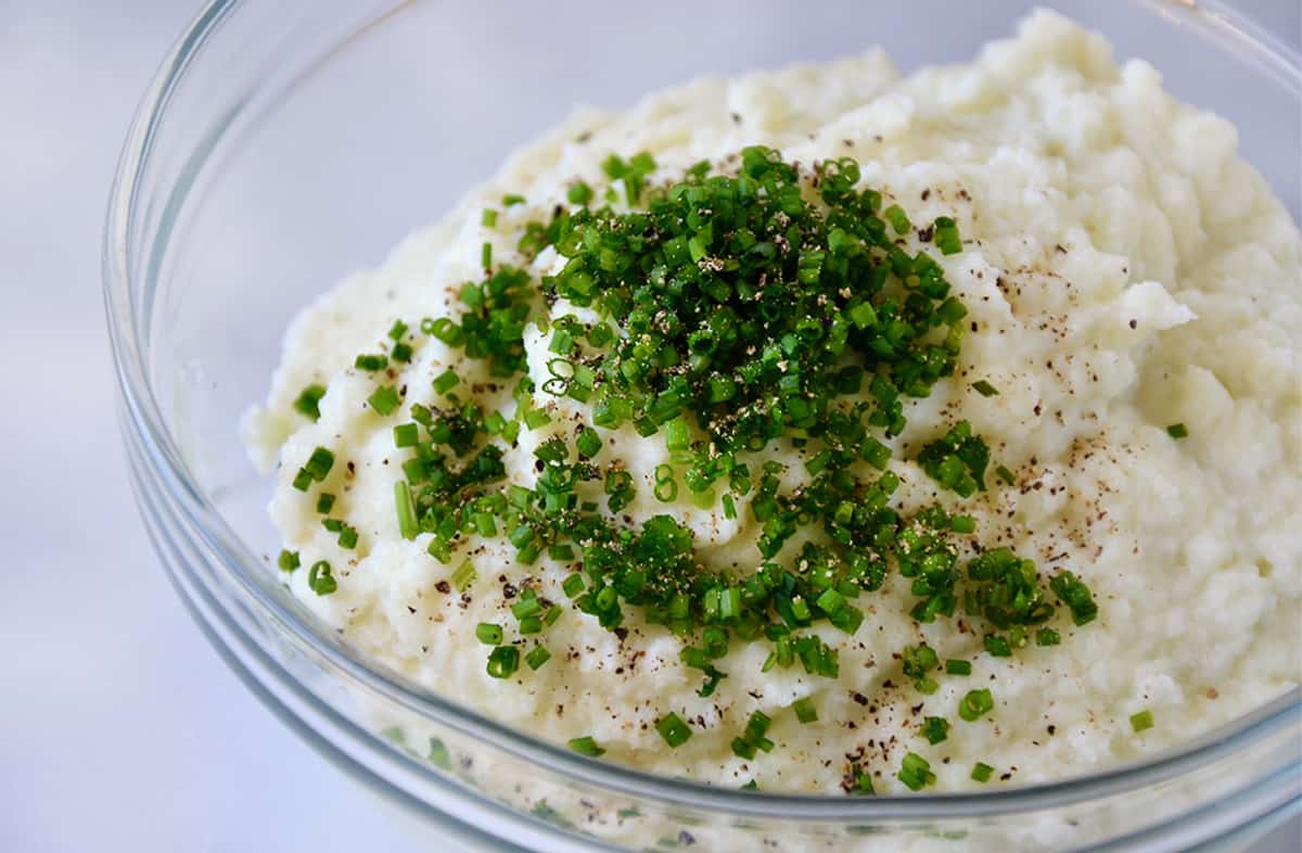 Mashed cauliflower, minced chives and black pepper in a glass mixing bowl.
