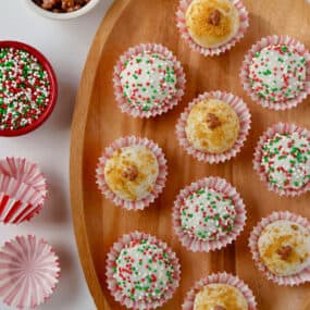 A wooden platter holding 10 chocolate- and sprinkle-covered gingerbread cookie balls in red-and-white striped wrappers. More wrappers and two small bowls of sprinkles sit beside the platter.