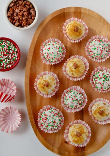 A wooden platter holding 10 chocolate- and sprinkle-covered gingerbread cookie balls in red-and-white striped wrappers. More wrappers and two small bowls of sprinkles sit beside the platter.