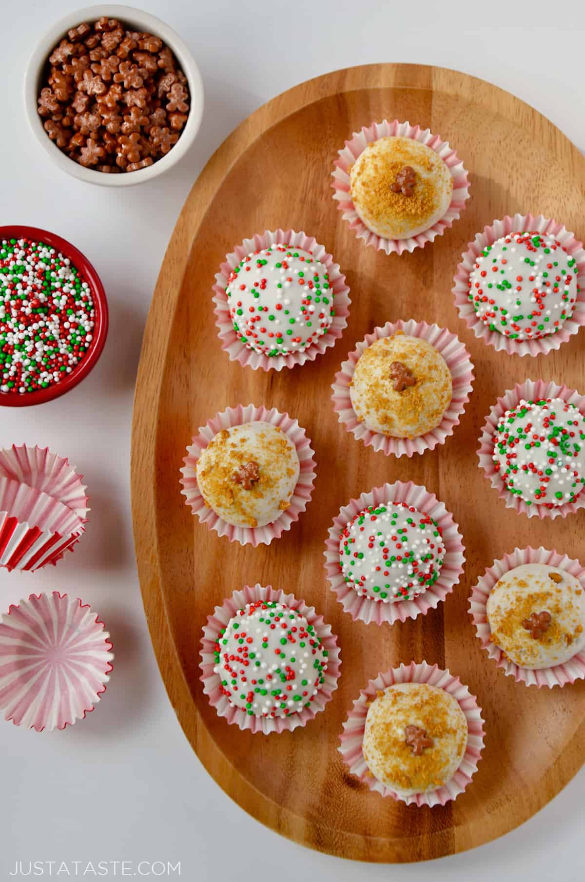 A wooden platter holding 10 chocolate- and sprinkle-covered gingerbread cookie balls in red-and-white striped wrappers. More wrappers and two small bowls of sprinkles sit beside the platter.