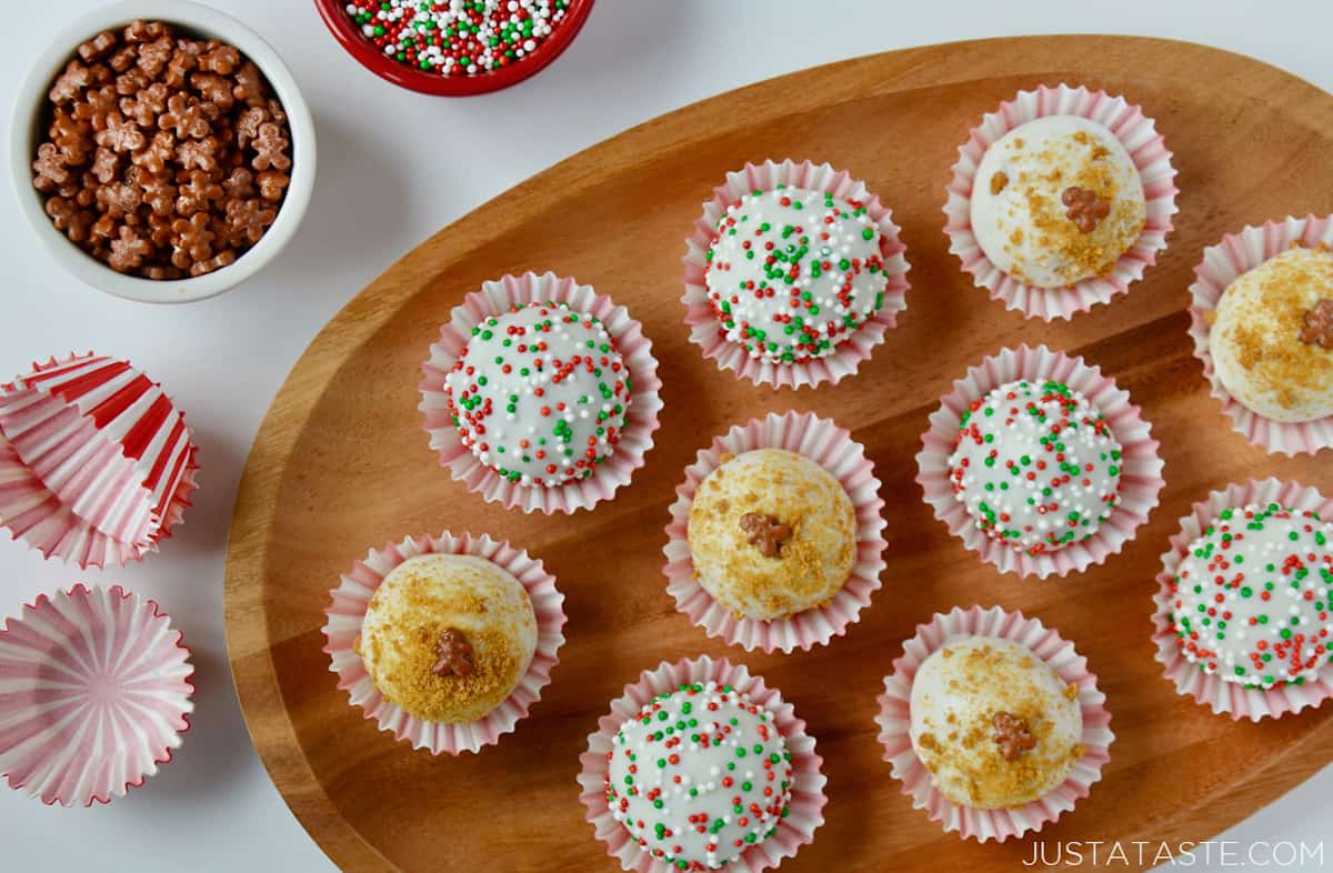 A wooden platter holding 10 white chocolate- and sprinkle-covered gingerbread cookie balls in red-and-white striped wrappers. More wrappers and two bowls of sprinkles sit beside the platter.