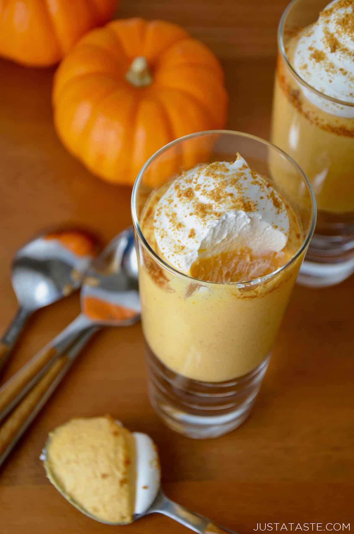 Two small serving glasses filled with no-bake pumpkin cheesecake and topped with whipped cream and gingersnap crumbs. Spoons and small pumpkins sit beside the glasses.