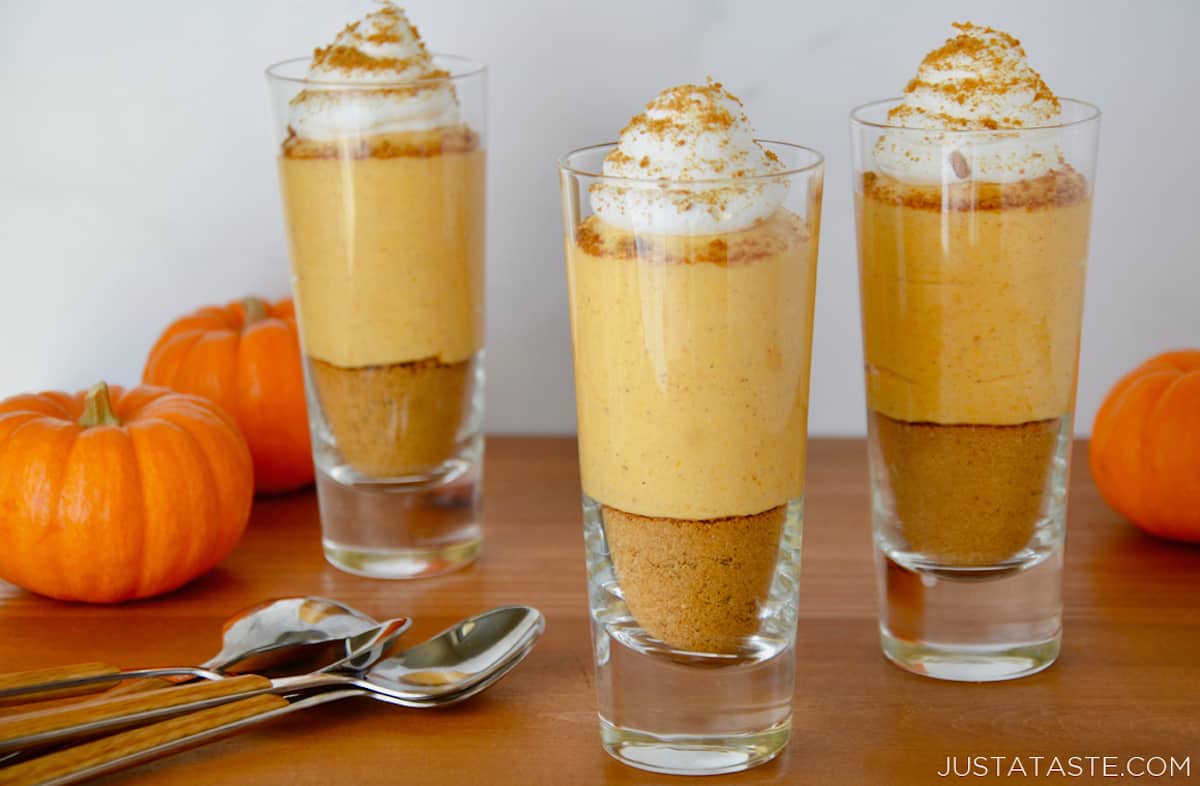 Three serving glasses filled with layers of cookie crumbs, no-bake pumpkin cheesecake and whipped cream. Spoons and three small pumpkins are beside the glasses.