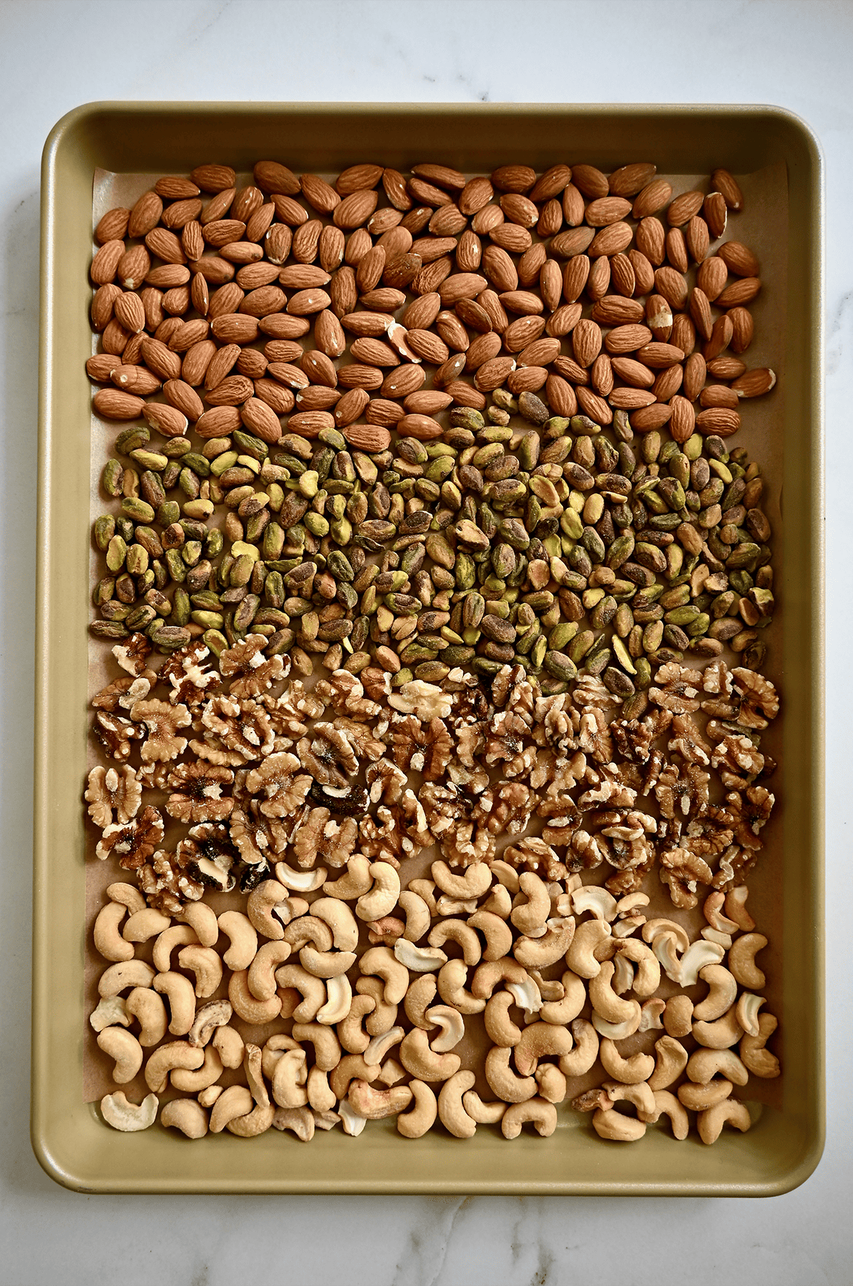 Rows of almonds, pistachios, walnuts and cashews on a parchment-lined baking sheet.