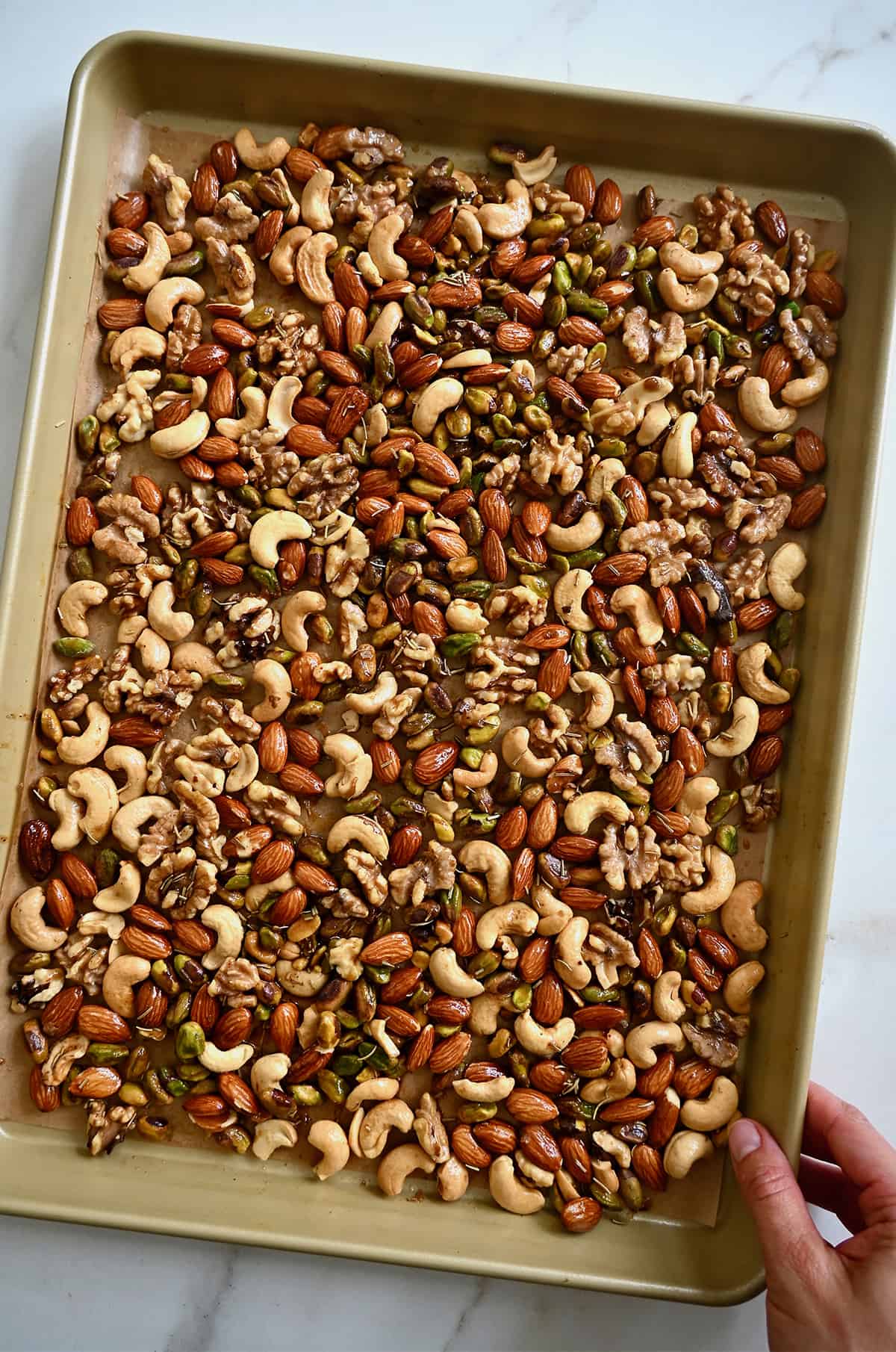 A person placing a baking sheet containing mixed nuts tossed with melted butter, spices and brown sugar onto a marble countertop.