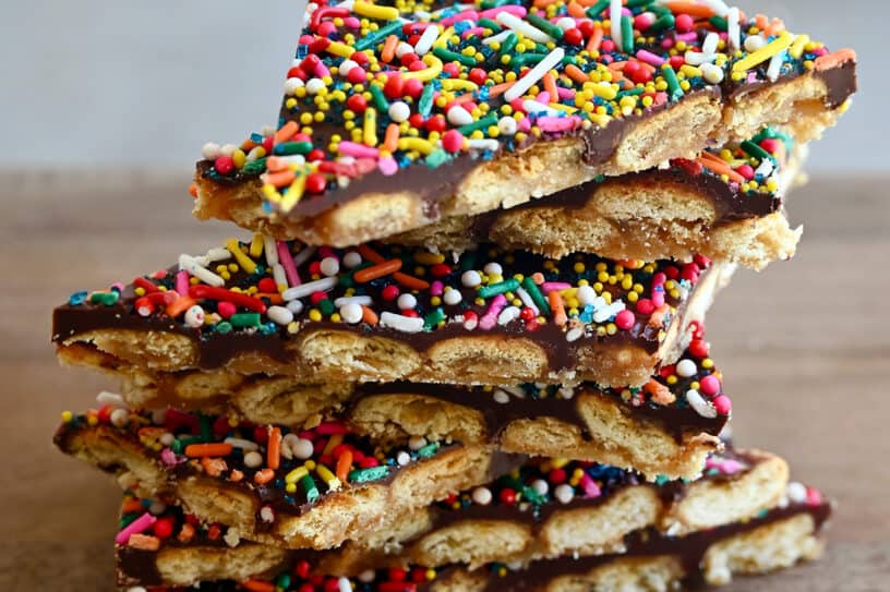 A stack of oyster cracker toffee bark with rainbow sprinkles on a wood cutting board.