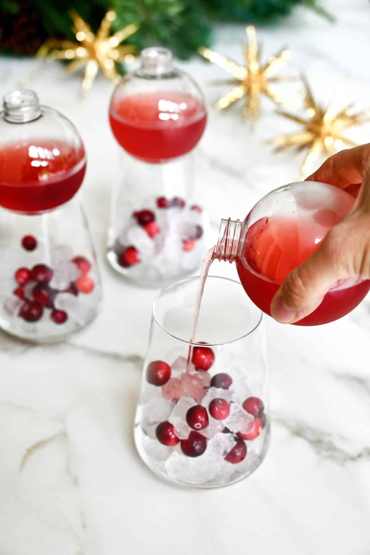 A hand holding a clear plastic ornament drinking ball pours Christmas margarita mix into a glass filled with ice and fresh cranberries.