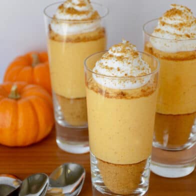 Three small glass cups containing no-bake pumpkin cheesecake parfaits that are topped with whipped cream. Spoons and small pumpkins sit beside the cups.