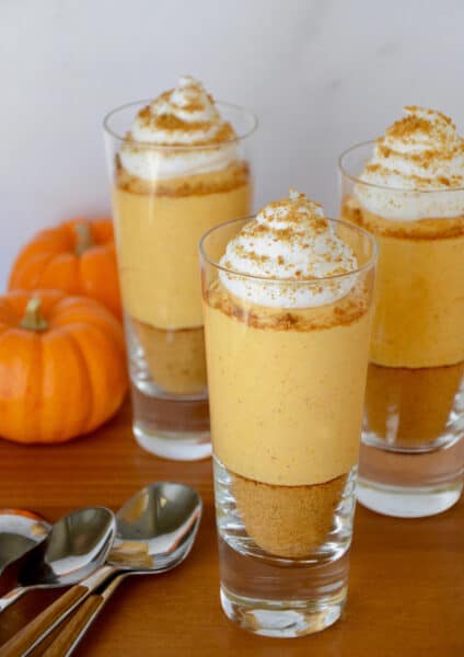 Three small glass cups containing no-bake pumpkin cheesecake parfaits that are topped with whipped cream. Spoons and small pumpkins sit beside the cups.