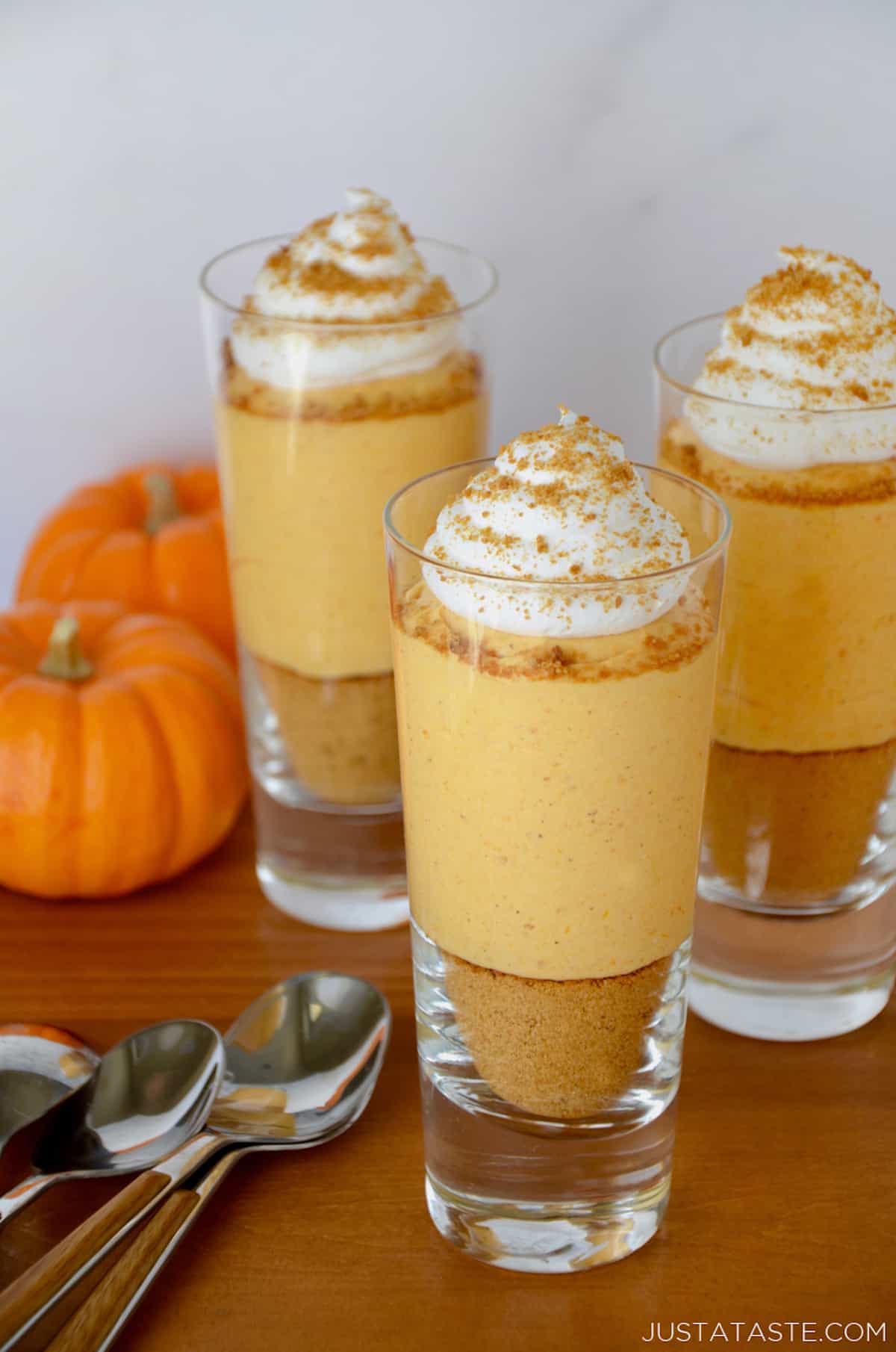 Three small glass cups containing no-bake pumpkin cheesecake parfaits that are topped with whipped cream. Spoons and small pumpkins sit beside the cups.