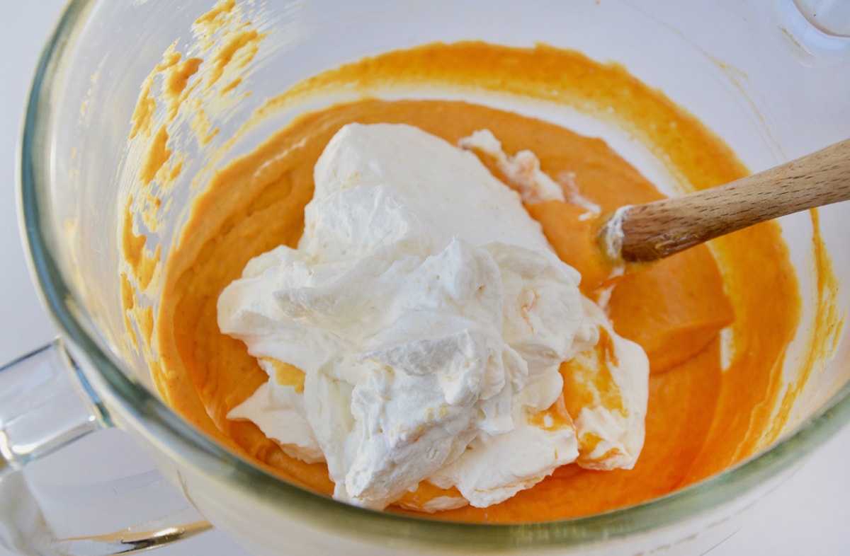Whipped cream on top of pumpkin filling in the bowl of a stand mixer. A spatula is propped up against the side of the bowl.