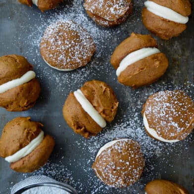 Pumpkin whoopie pies on a baking sheet that are dusted with powdered sugar. A small fine mesh strainer with more powdered sugar sits off to the side.