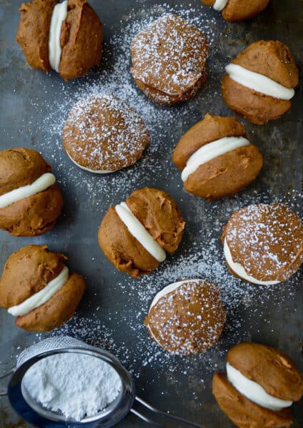 Pumpkin whoopie pies on a baking sheet that are dusted with powdered sugar. A small fine mesh strainer with more powdered sugar sits off to the side.