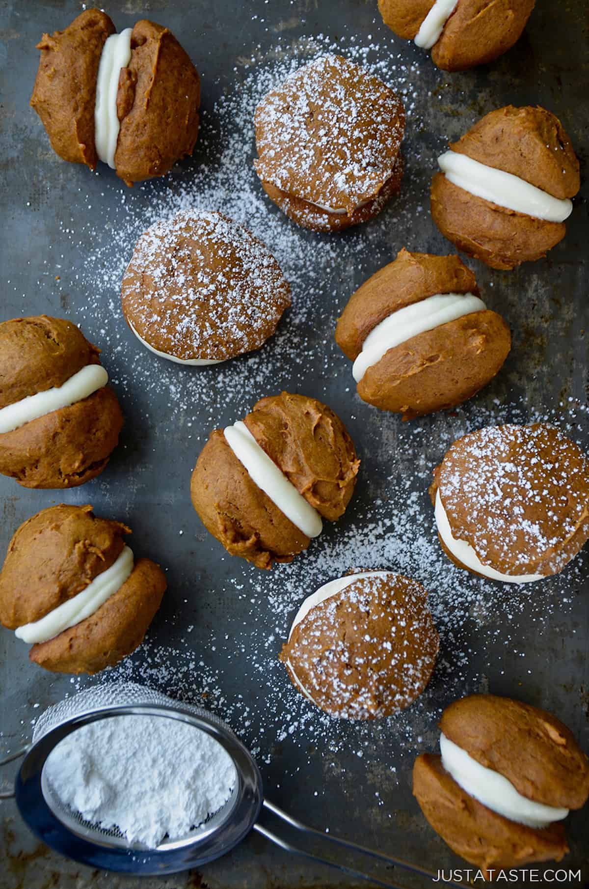 Pumpkin whoopie pies on a baking sheet that are dusted with powdered sugar. A small fine mesh strainer with more powdered sugar sits off to the side.