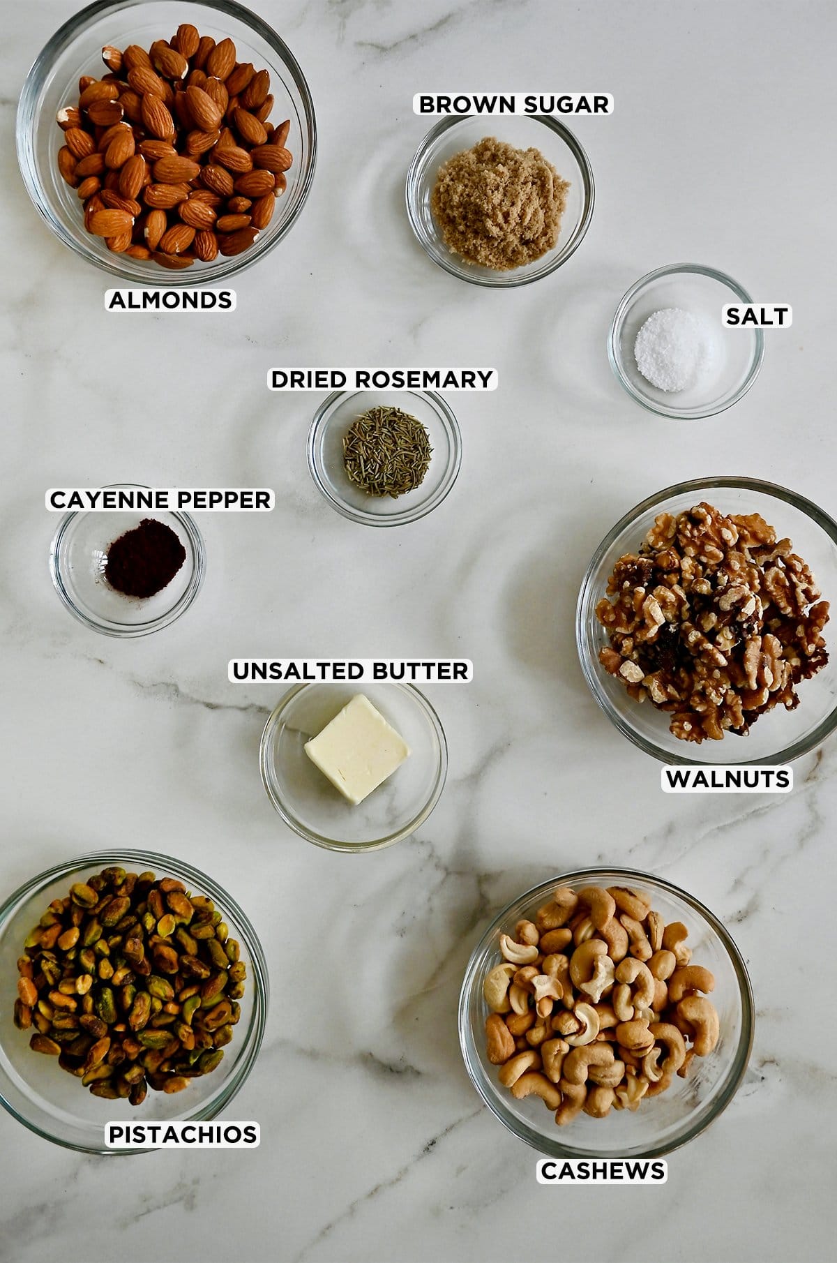 Small glass bowls containing cashews, pistachios, almonds, walnuts, butter, dried rosemary, cayenne pepper, salt and brown sugar on a marble countertop.