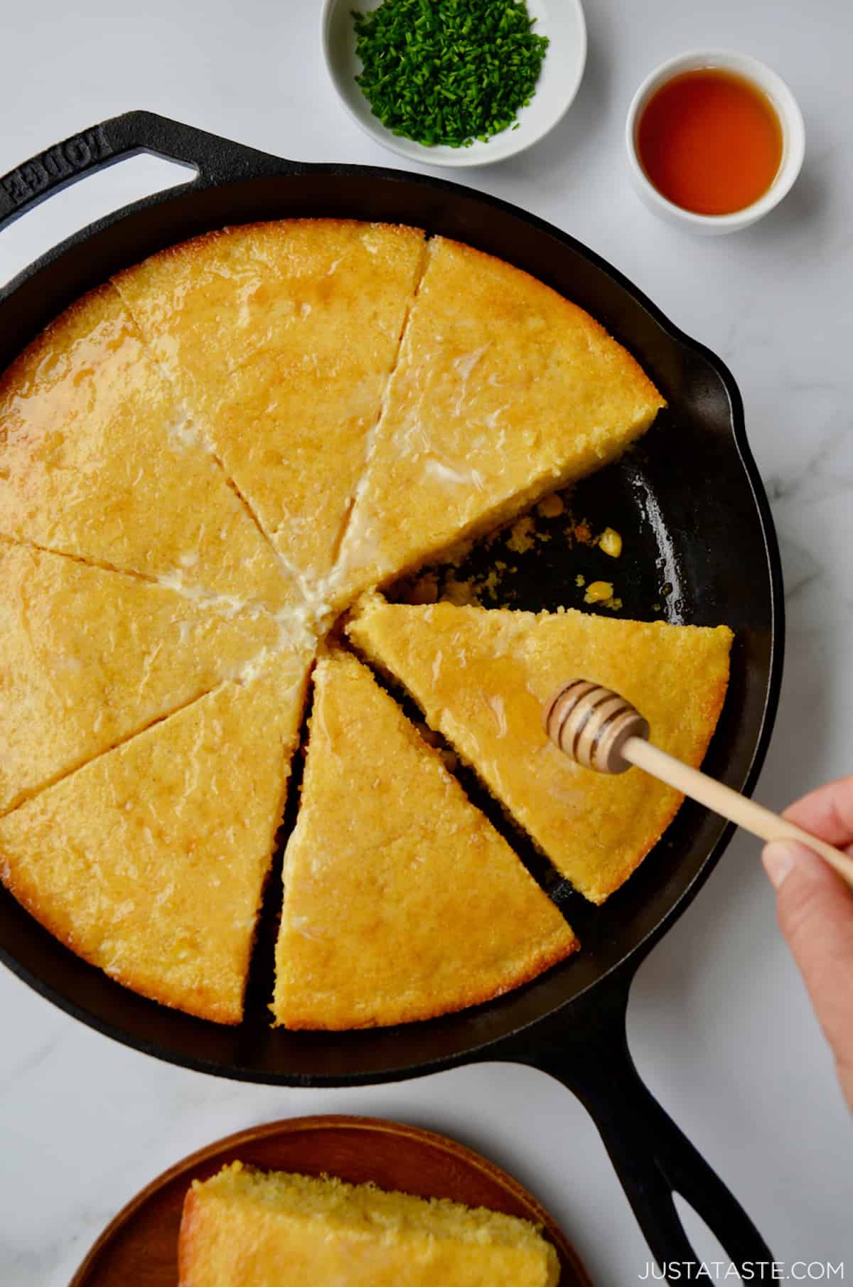 A person drizzling honey over a sliced cornbread in a cast iron pan. Surrounding the pan is a plate with a slice of cornbread on it, a small bowl with minced chives and a small bowl of honey.