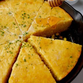 Honey being drizzled over sliced cornbread in a cast iron pan. The cornbread is topped with butter and minced chives and small bowls of more chives and honey are beside the skillet.