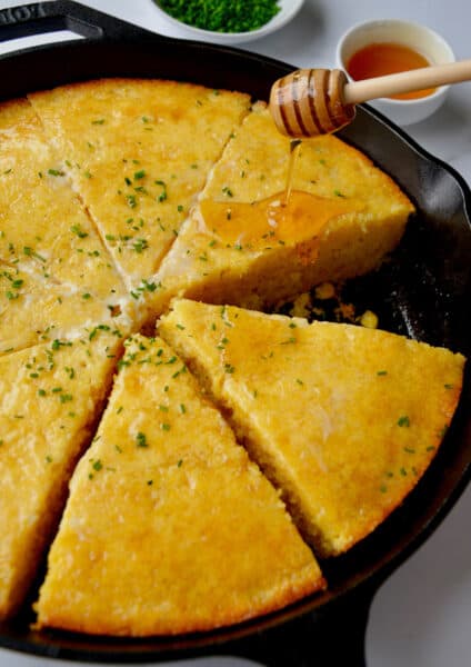 Honey being drizzled over sliced cornbread in a cast iron pan. The cornbread is topped with butter and minced chives and small bowls of more chives and honey are beside the skillet.